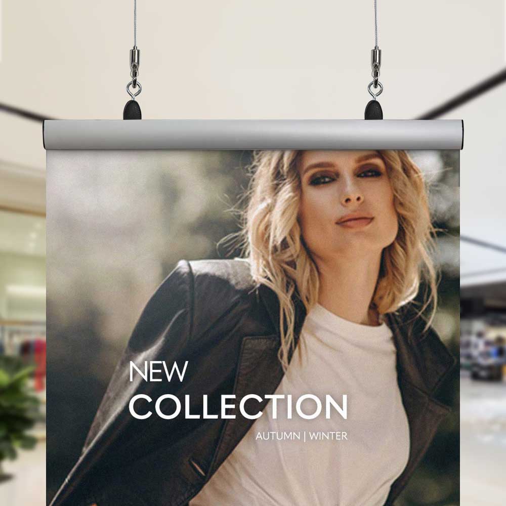 Image shows a poster clamp sign being hung via two cables and safe hooks from the top left and right corners. Sign shows a woman wearing a coat and reads 'New collection autumn winter' in a shop.