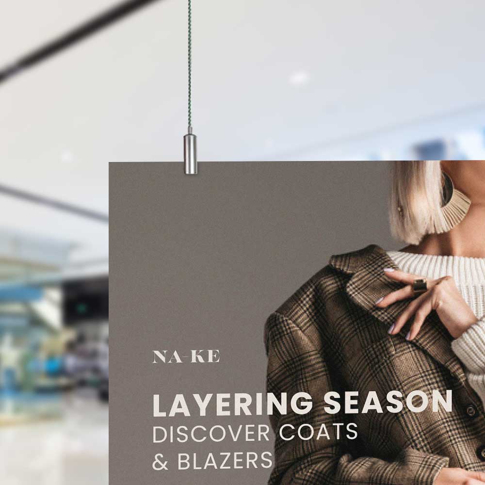 Image shows the top left corner panel sign being hung via cables and a panel gripper. Sign shows the shoulder of a woman touching the collar of a coat and reads 'na-ke layering season discover coats & blazers