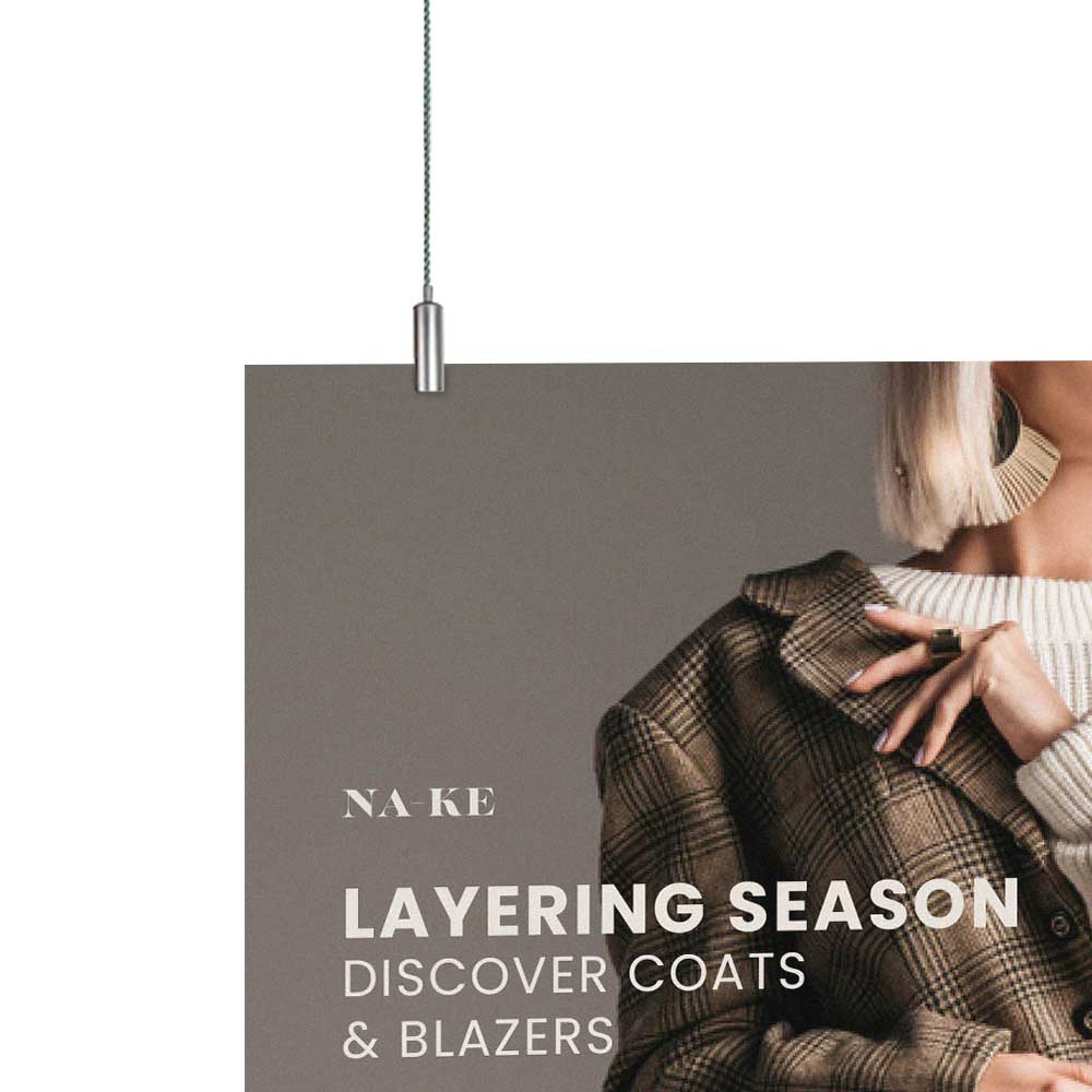 Image shows the top left corner panel sign being hung via cables and a panel gripper. Sign shows the shoulder of a woman touching the collar of a coat and reads 'na-ke layering season discover coats & blazers